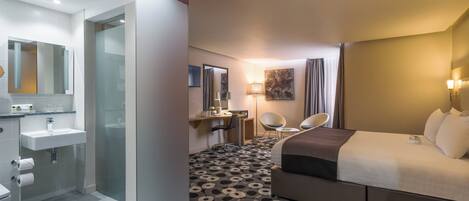 A hotel room with a bed, a desk, a chair, a bathroom with a sink, and a mirror.