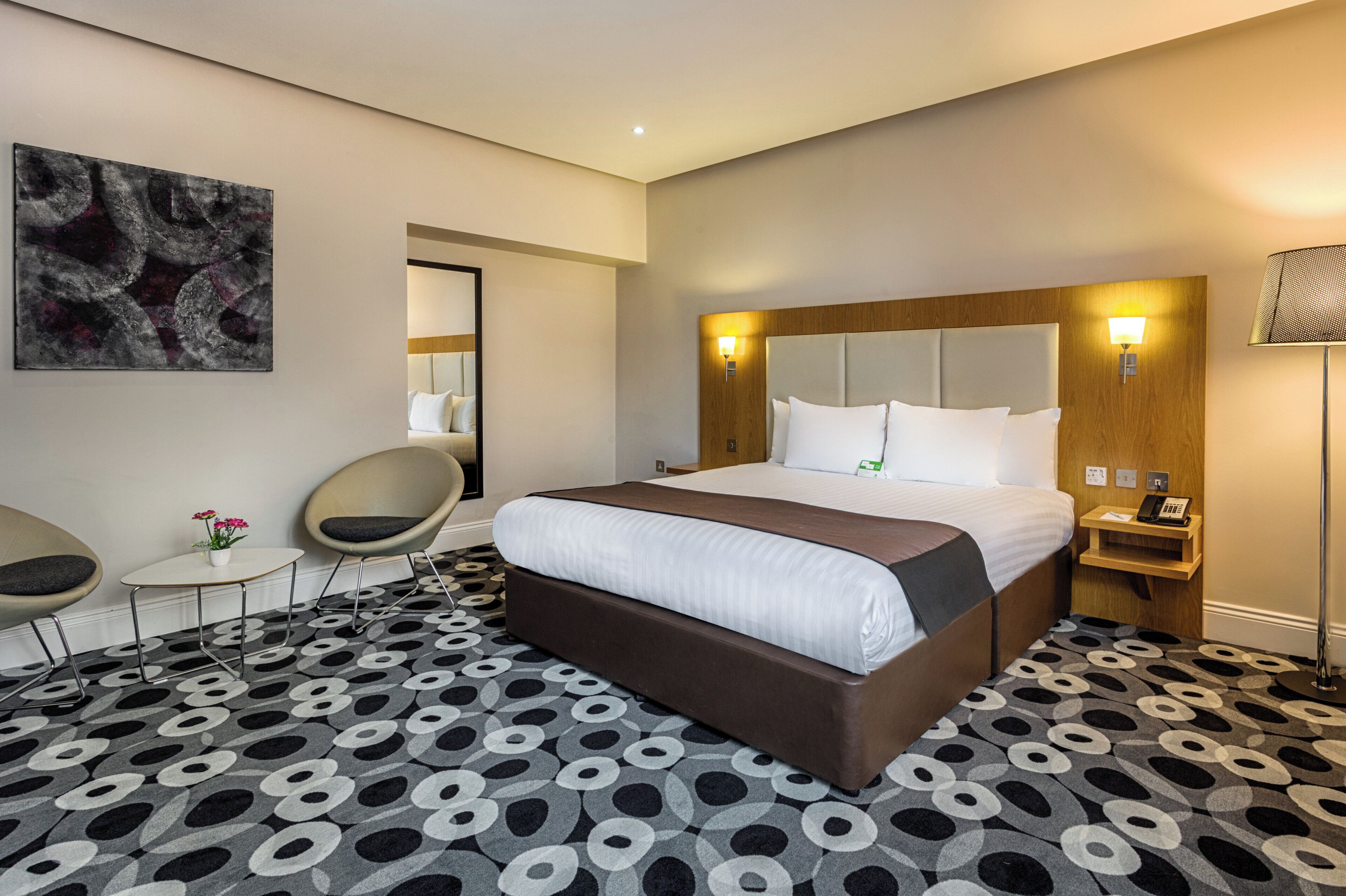 A hotel room with a large bed, a desk, two chairs, a painting on the wall, and a patterned carpet.