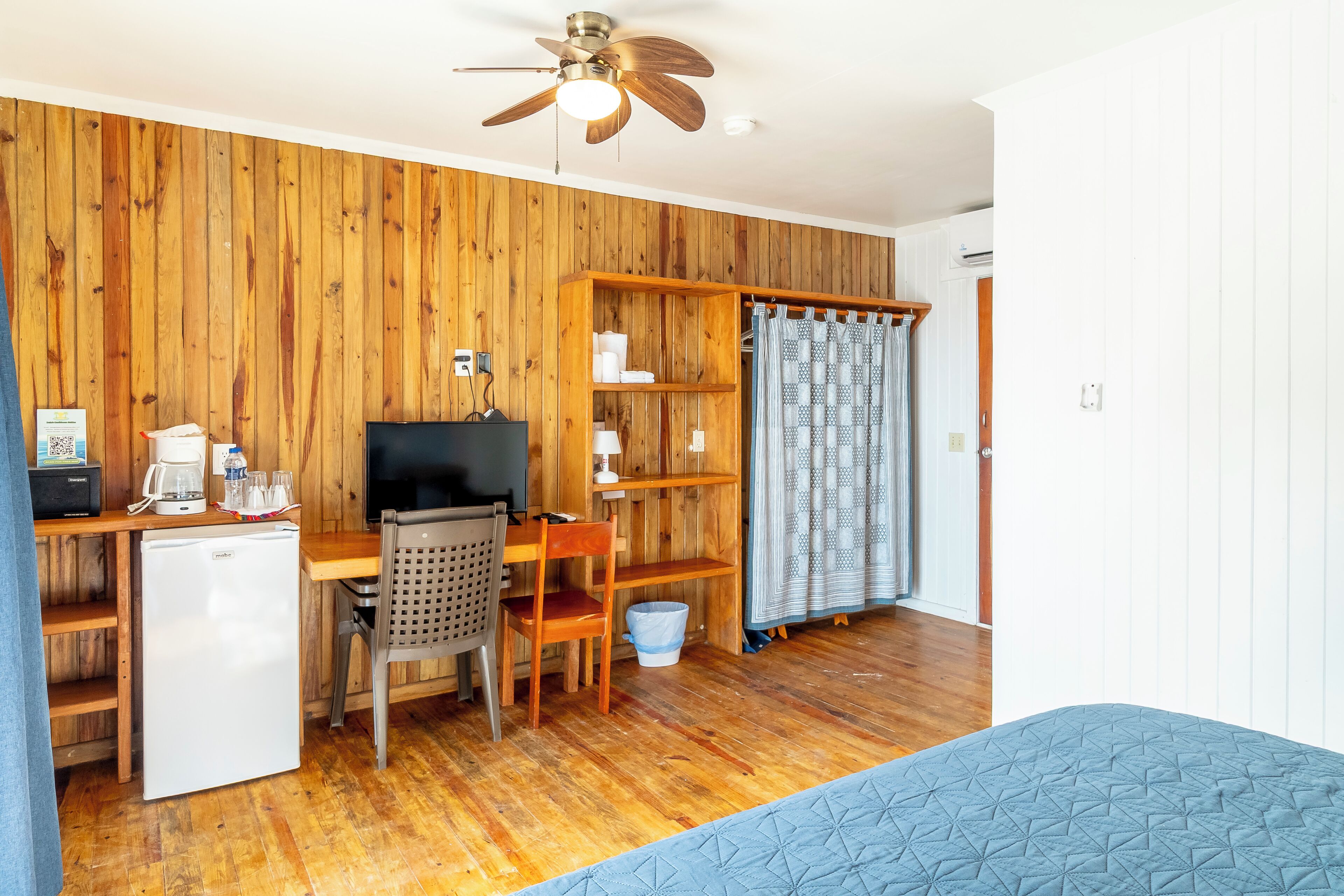 A room with a wooden wall, a ceiling fan, a desk with a computer, a chair, a small fridge, and a bed with a blue quilt.