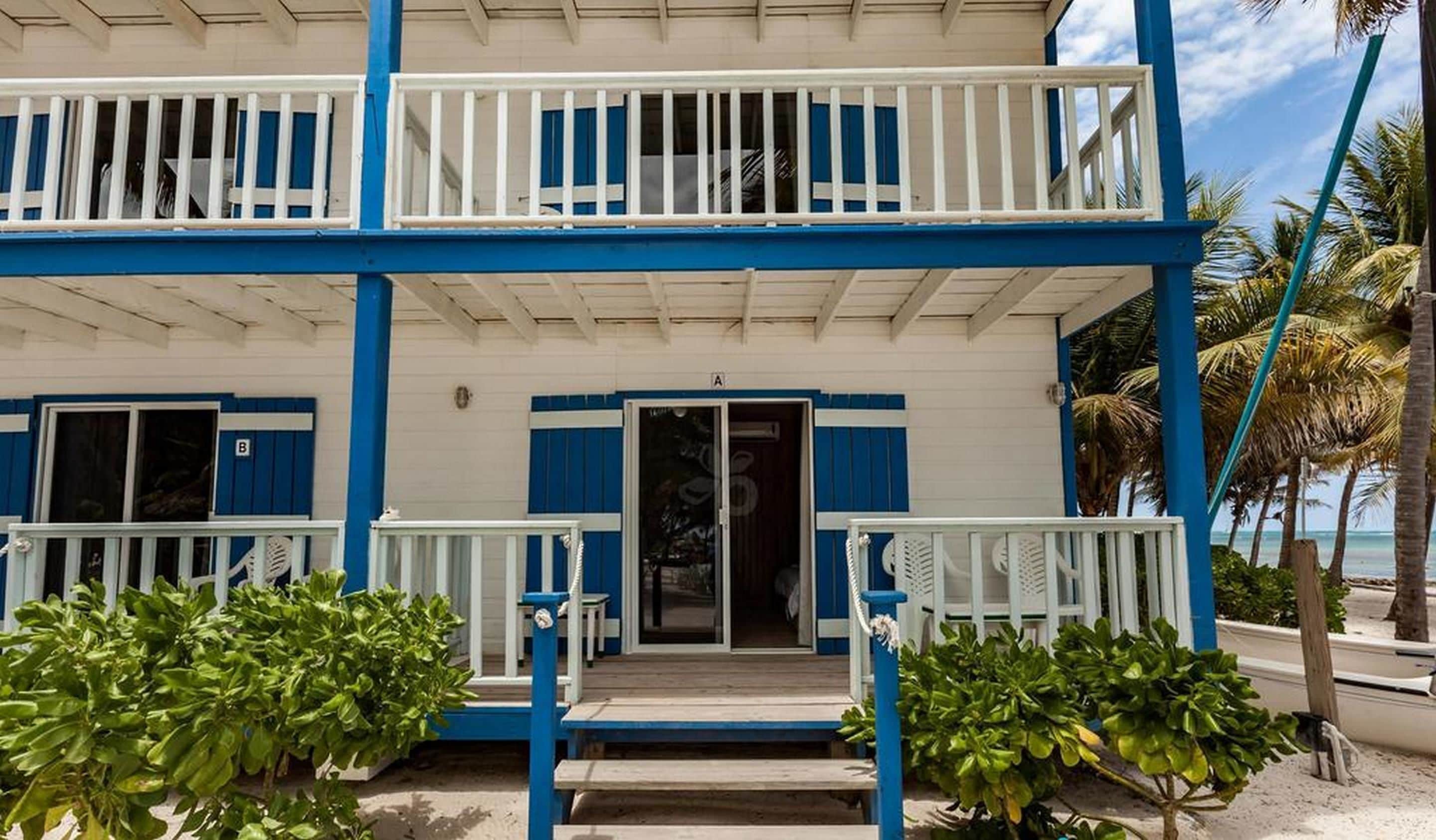 A two-story beachfront building with balconies, a covered porch, and a sandy ground.