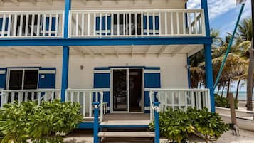 Un edificio frente a la playa de dos pisos, con balcones, un porche cubierto y suelo arenoso.