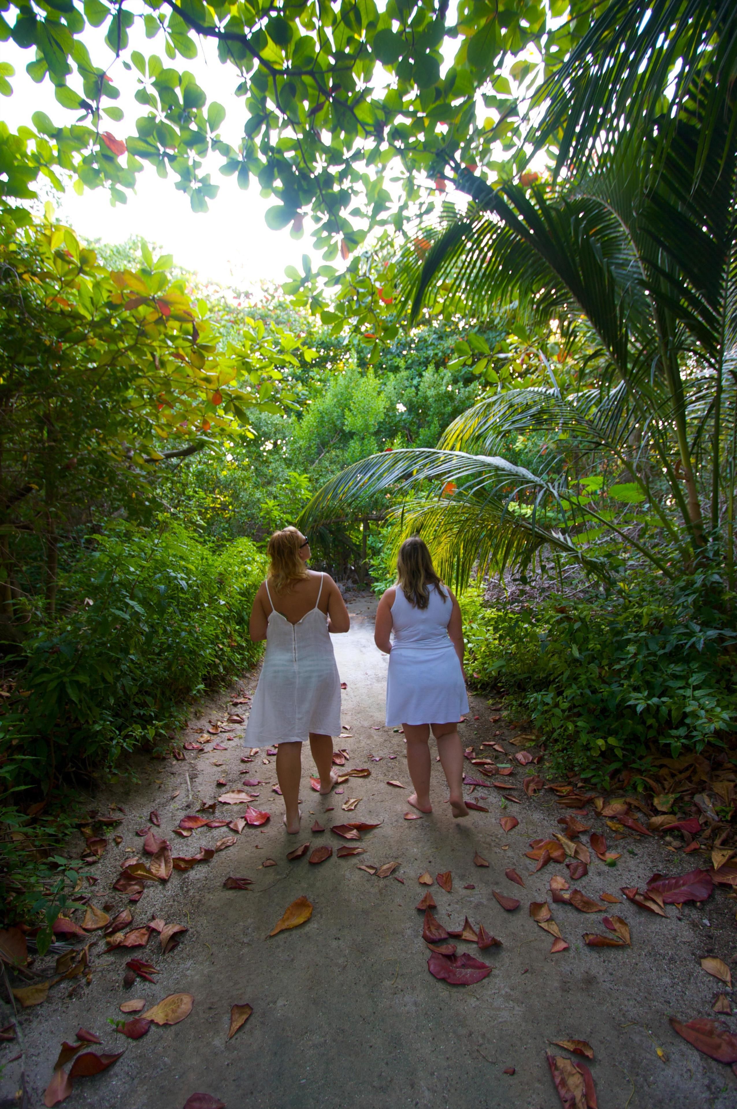 Duas mulheres caminham por um caminho rodeado por vegetação exuberante e palmeiras.
