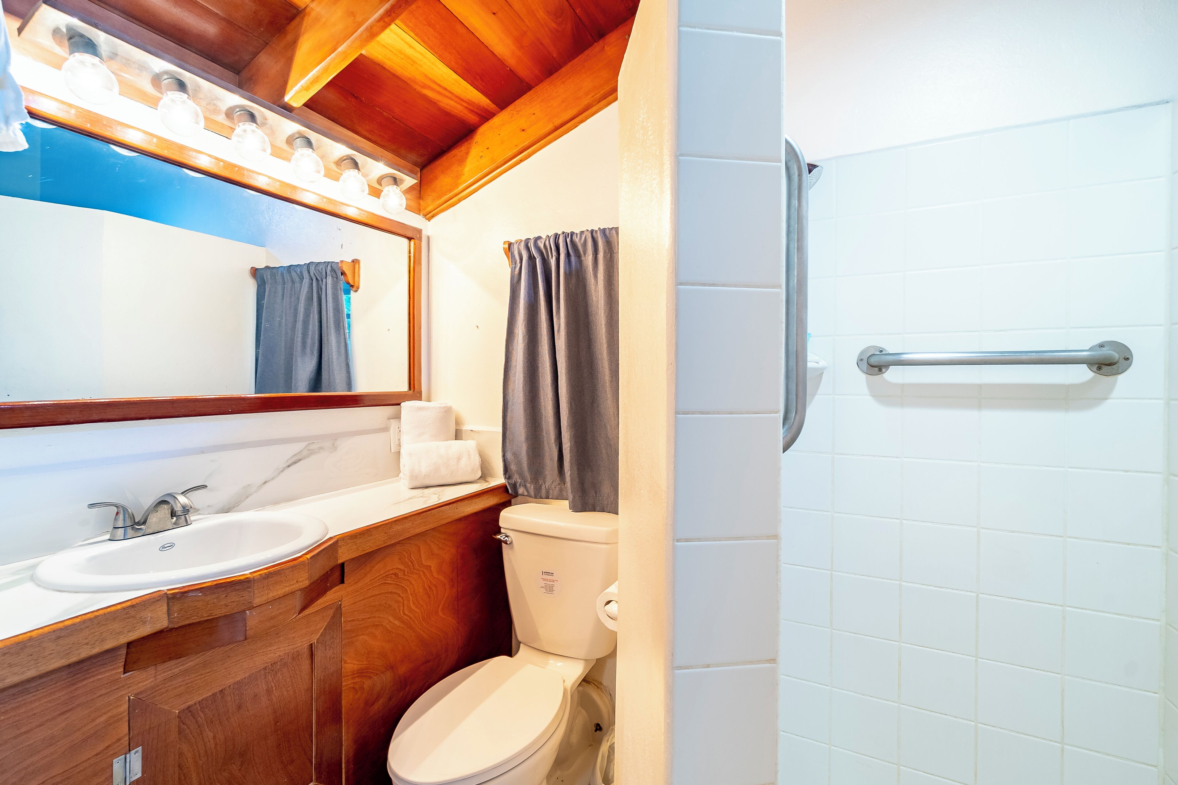 Un baño con tocador de madera, lavamanos blanco, inodoro y zona de ducha con barra de apoyo.