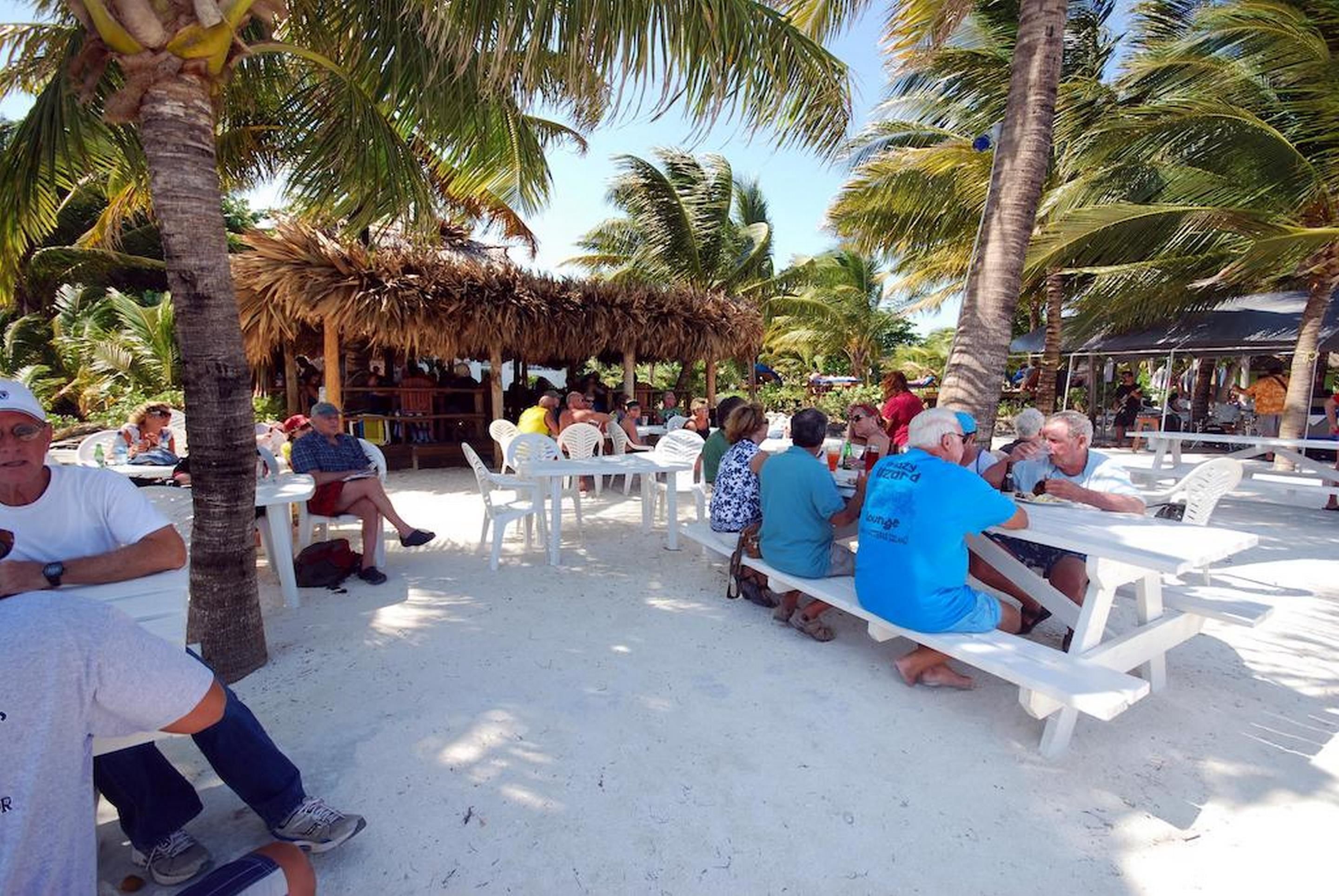 A beachside dining area with white tables and benches, surrounded by palm trees and a thatched-roof structure.