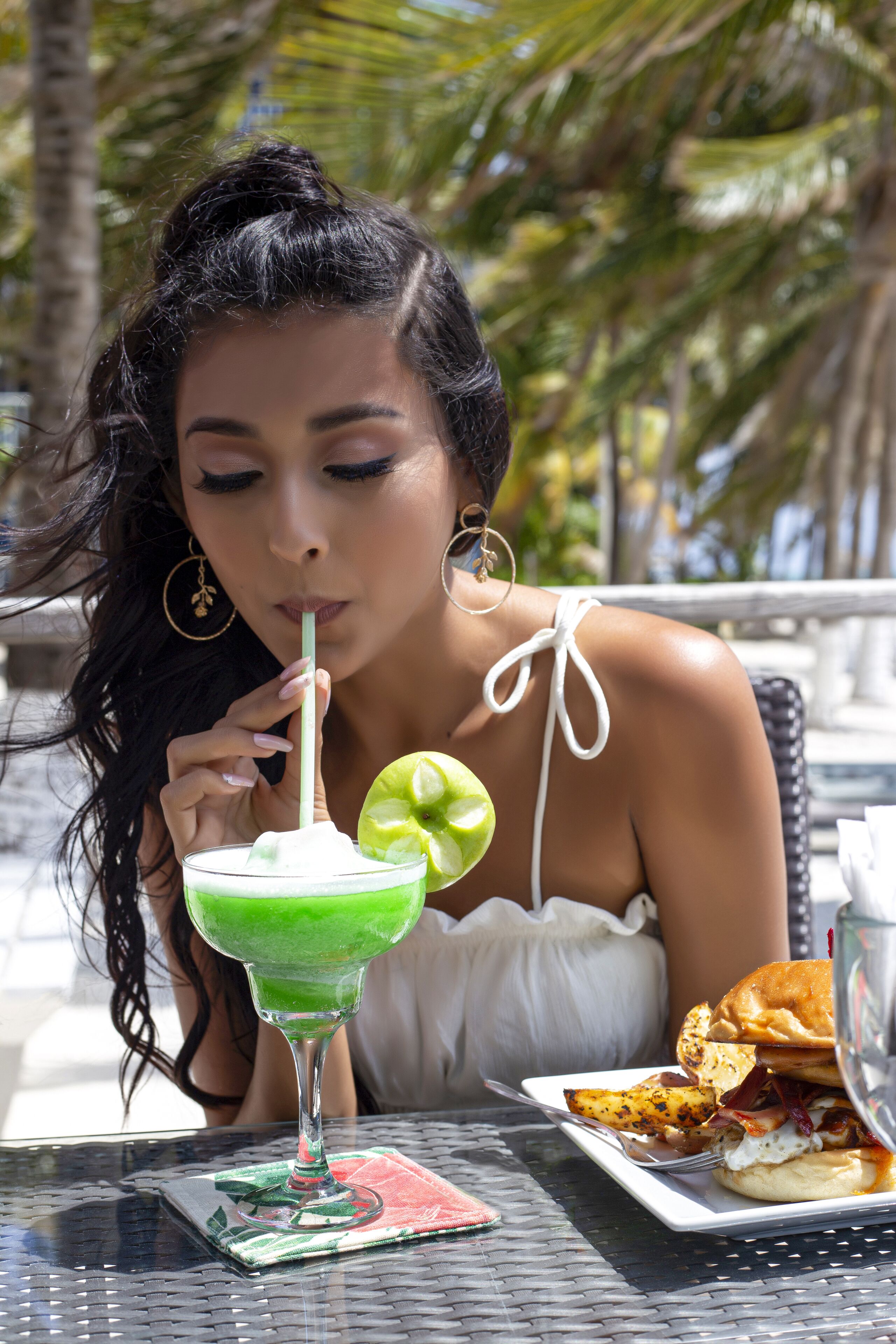 A person sitting at an outdoor table with a green drink, a plate with food, and a smartphone on the table.