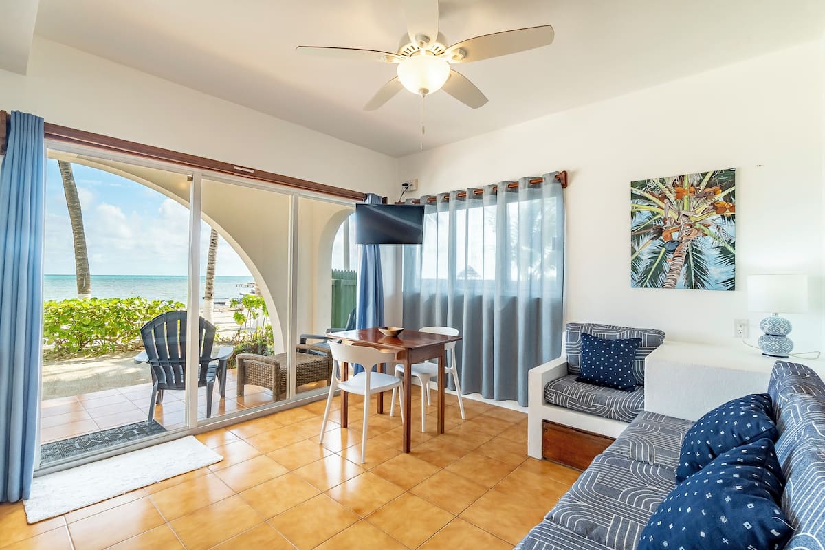 A room with a large sliding glass door leading to a balcony with a view of the ocean. There is a ceiling fan, a dining table with chairs, and a sofa with patterned cushions.