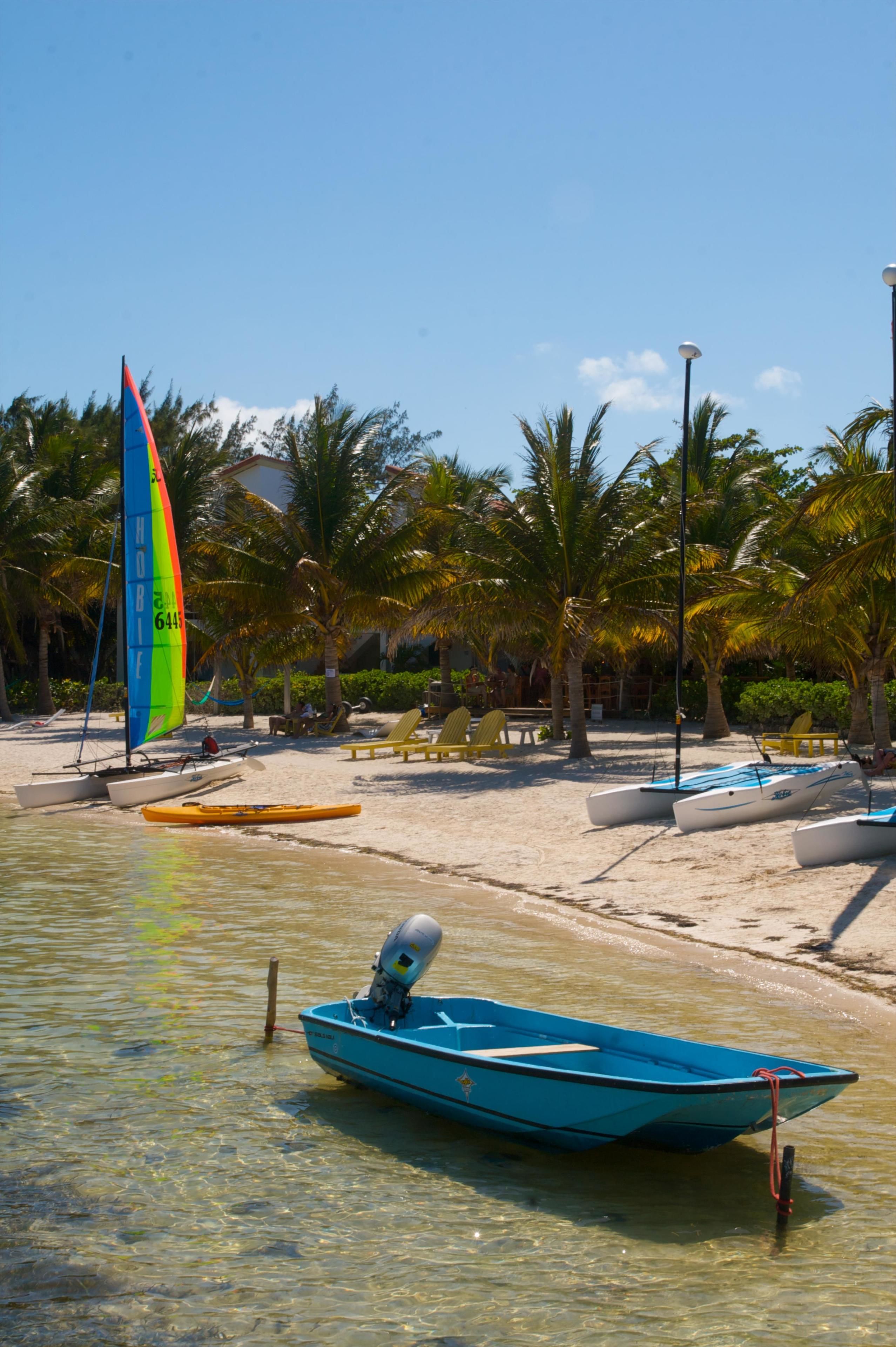 Una playa con un velero, un bote y un barco a motor. Hay palmeras y sillas de playa.