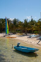 Una playa con un velero, un bote y un barco a motor. Hay palmeras y sillas de playa.
