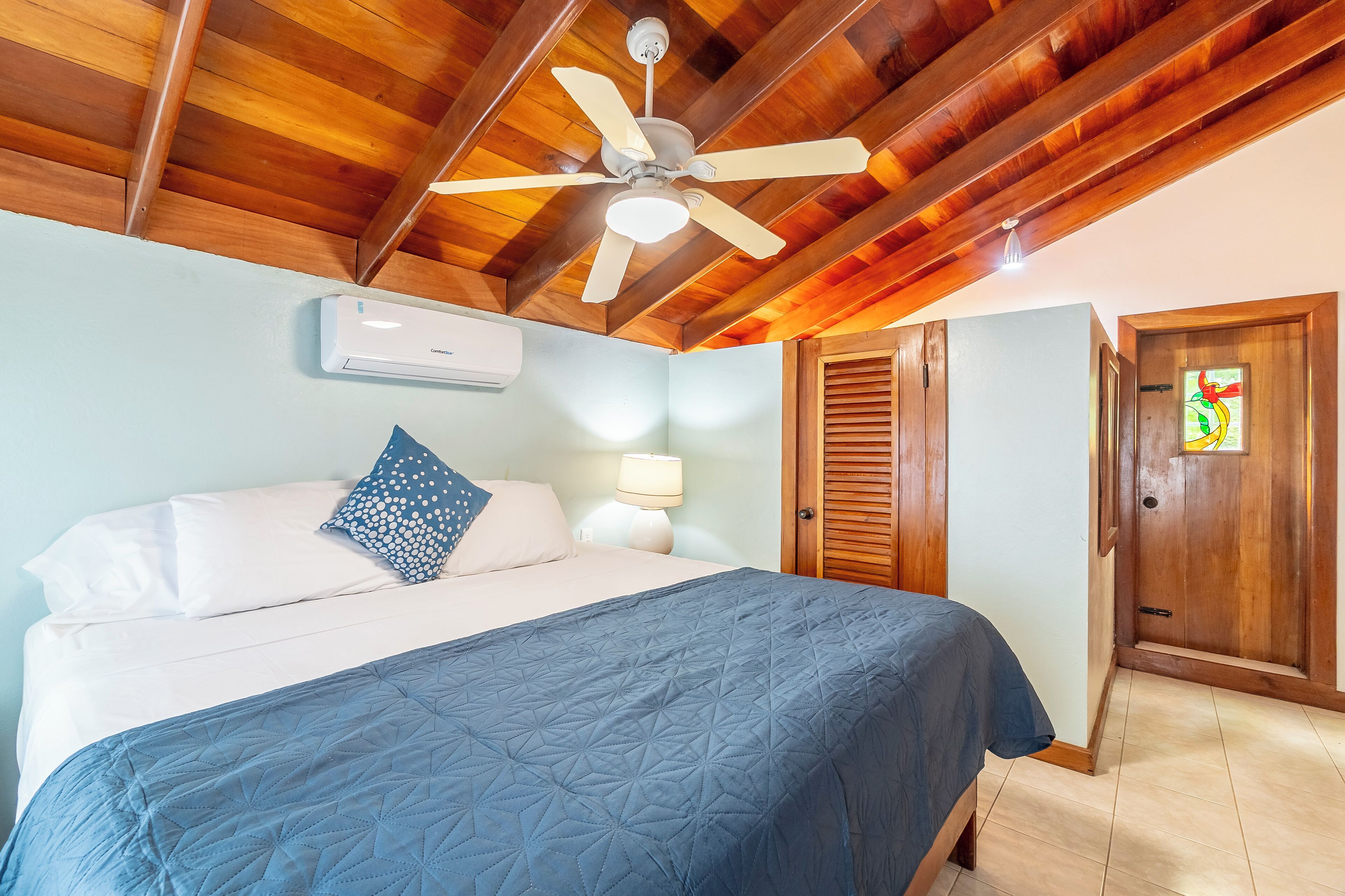 Un dormitorio con una cama, ventilador de techo, aire acondicionado de pared, lámpara de mesita de noche y puerta de madera.