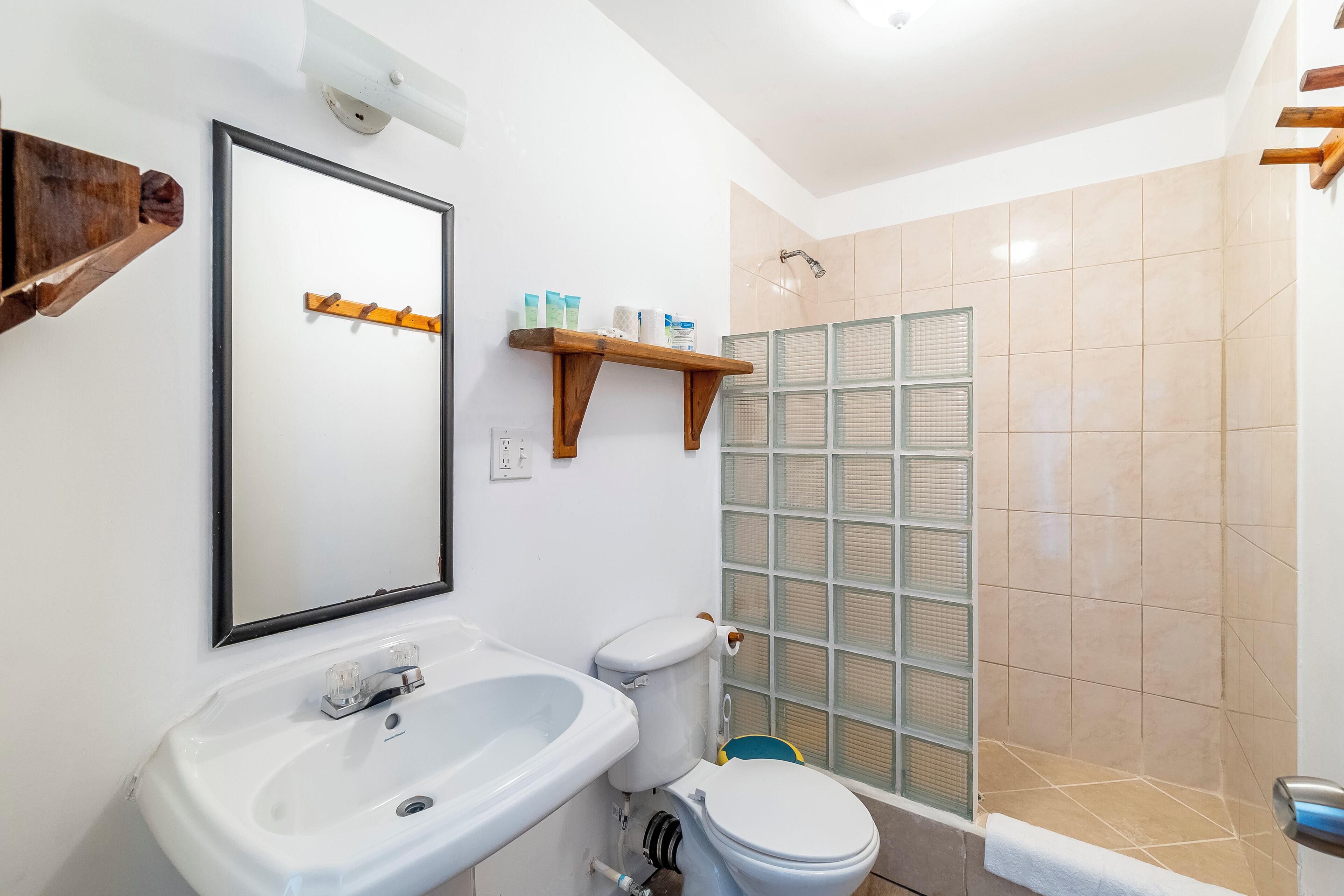 A bathroom with a white sink, toilet, and shower. There are wooden shelves above the toilet and a mirror on the wall.
