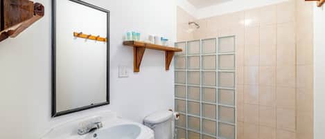 A bathroom with a white sink, toilet, and shower. There are wooden shelves above the toilet and a mirror on the wall.