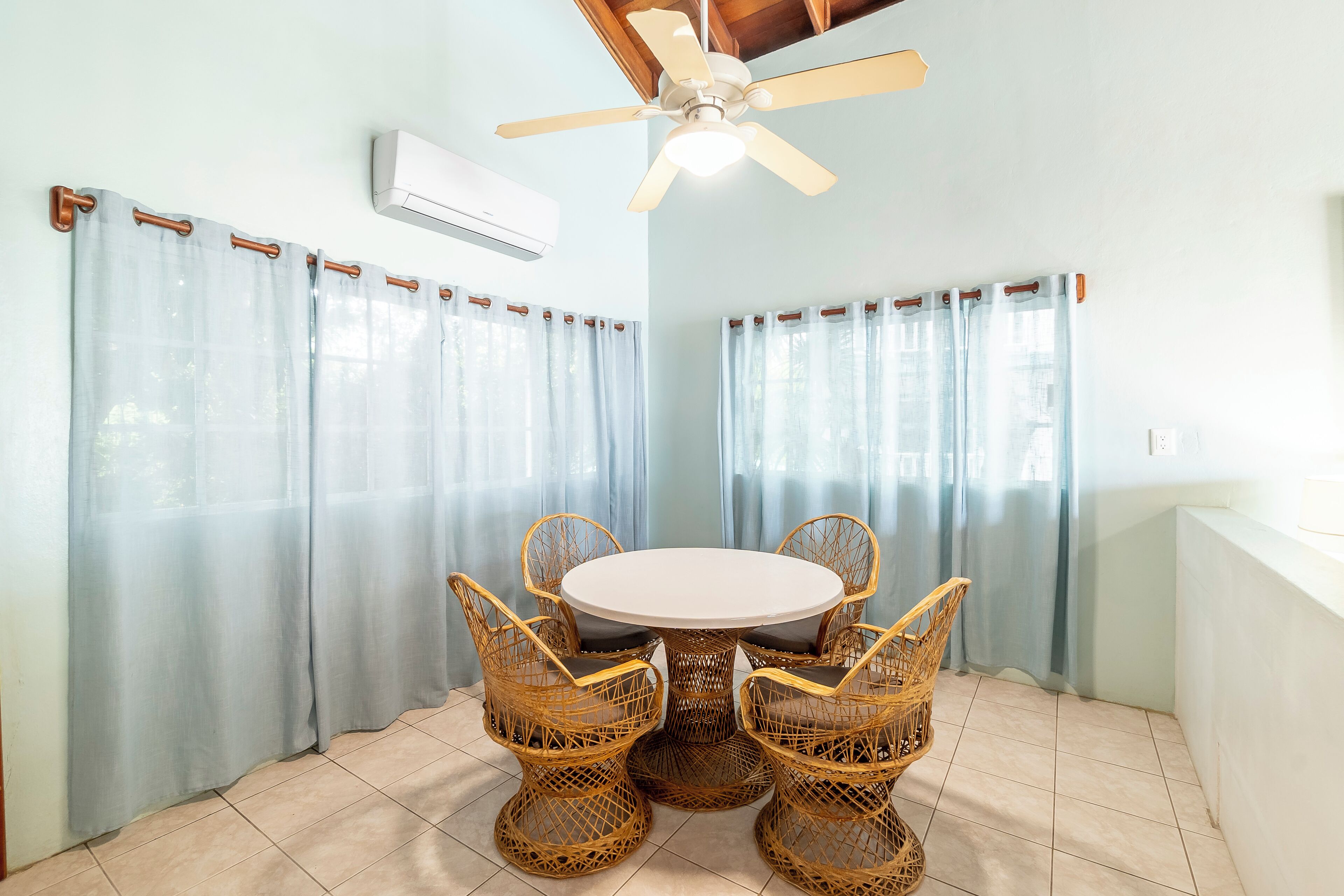 A dining area with a round table and four wicker chairs, light blue curtains, and a ceiling fan.
