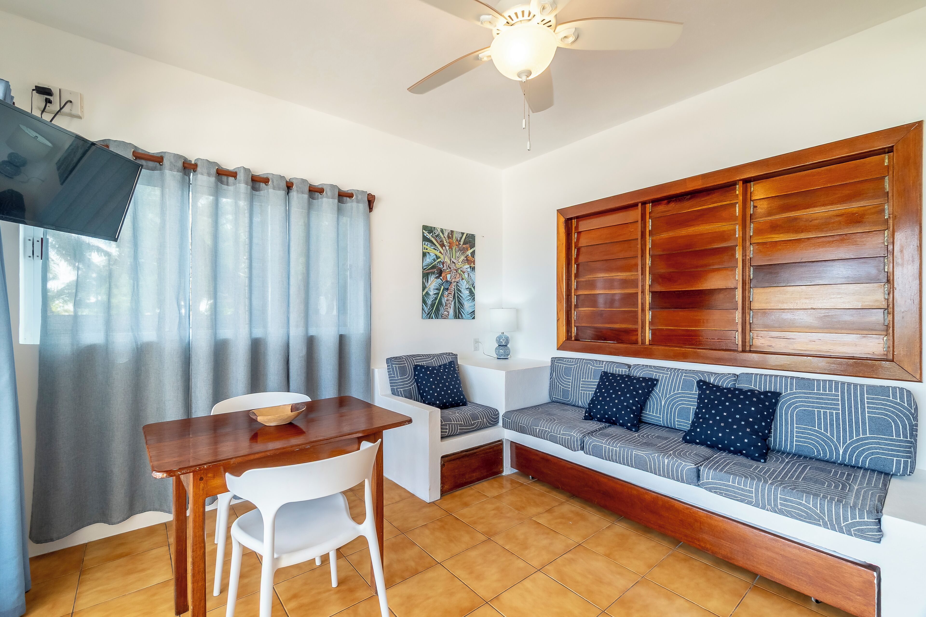 Un espacio habitable compacto con una mesa de madera, sillas blancas, un sofá con cojines estampados y un ventilador de techo.