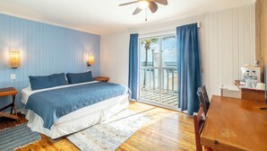 Un dormitorio con cama, suelo de madera, ventilador de techo y vista al océano a través de una puerta corredera de vidrio.