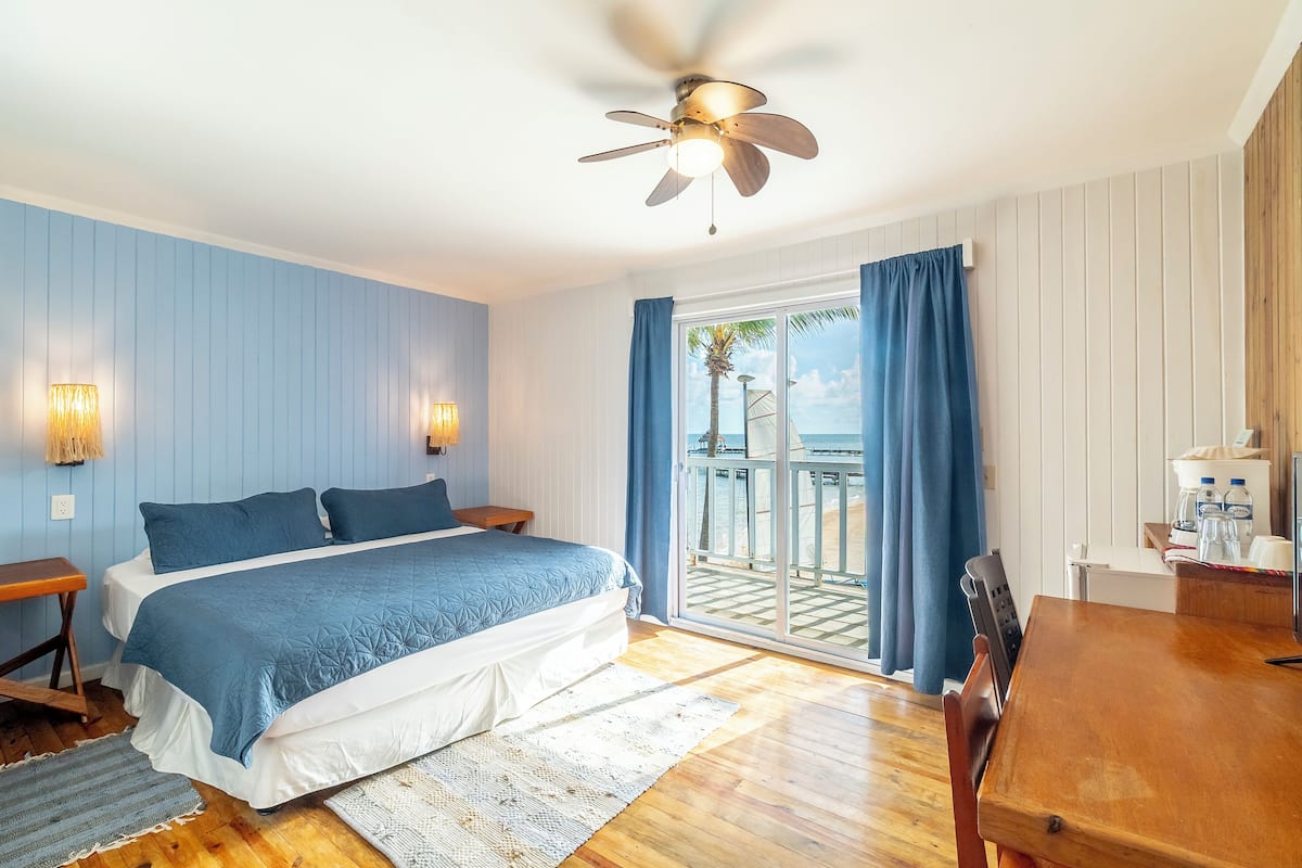 A bedroom with a bed, wooden floor, ceiling fan, and a view of the ocean through a sliding glass door.