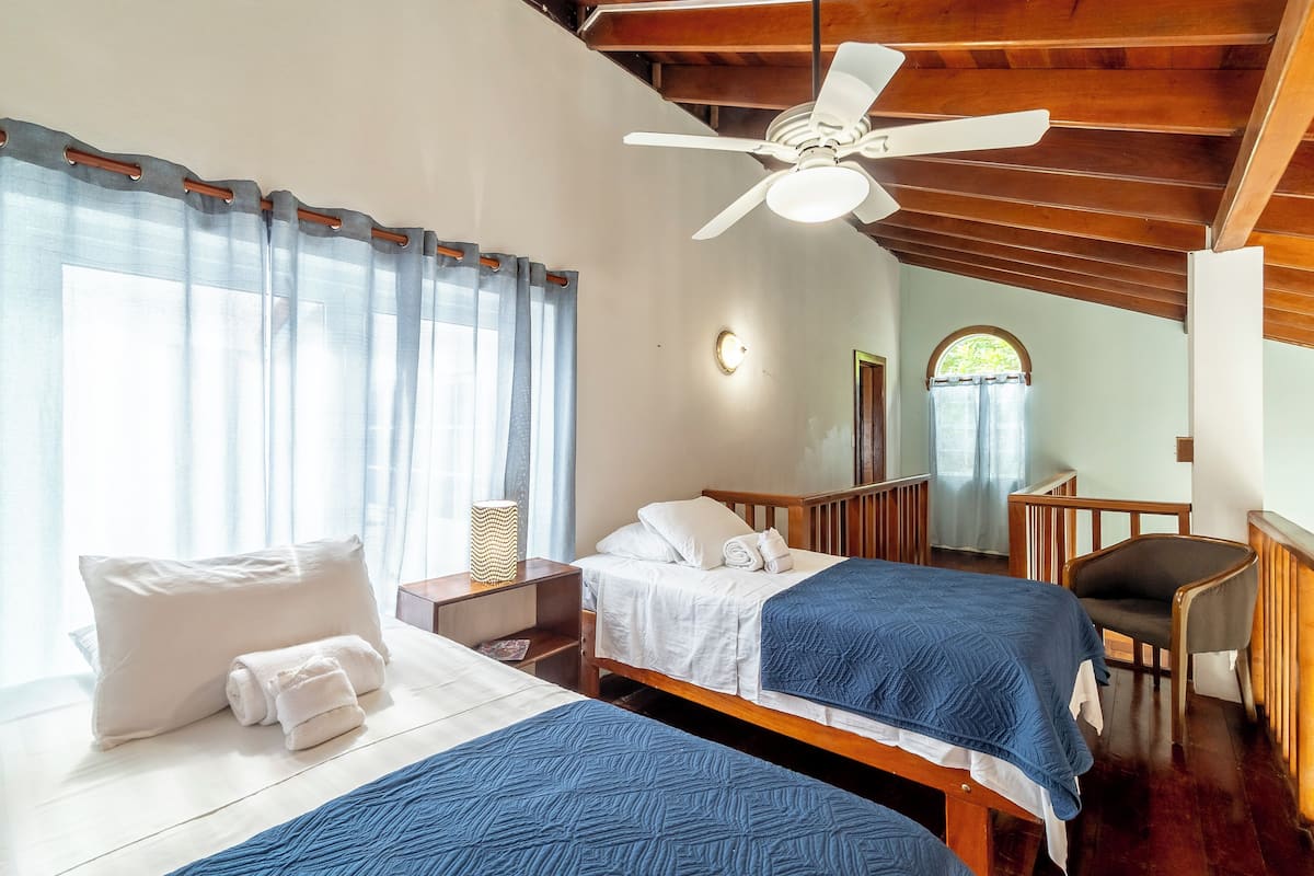 A hotel room with two beds, a ceiling fan, a nightstand, a chair, and a window with curtains.