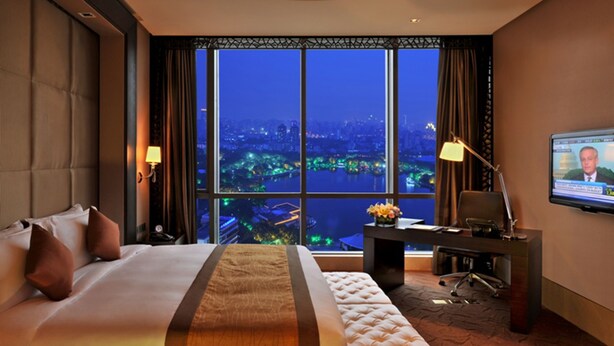 A hotel room with a large bed, a desk, a chair, a TV, and a view of a cityscape at night. - Guoman Hotel Shanghai (Shanghai)