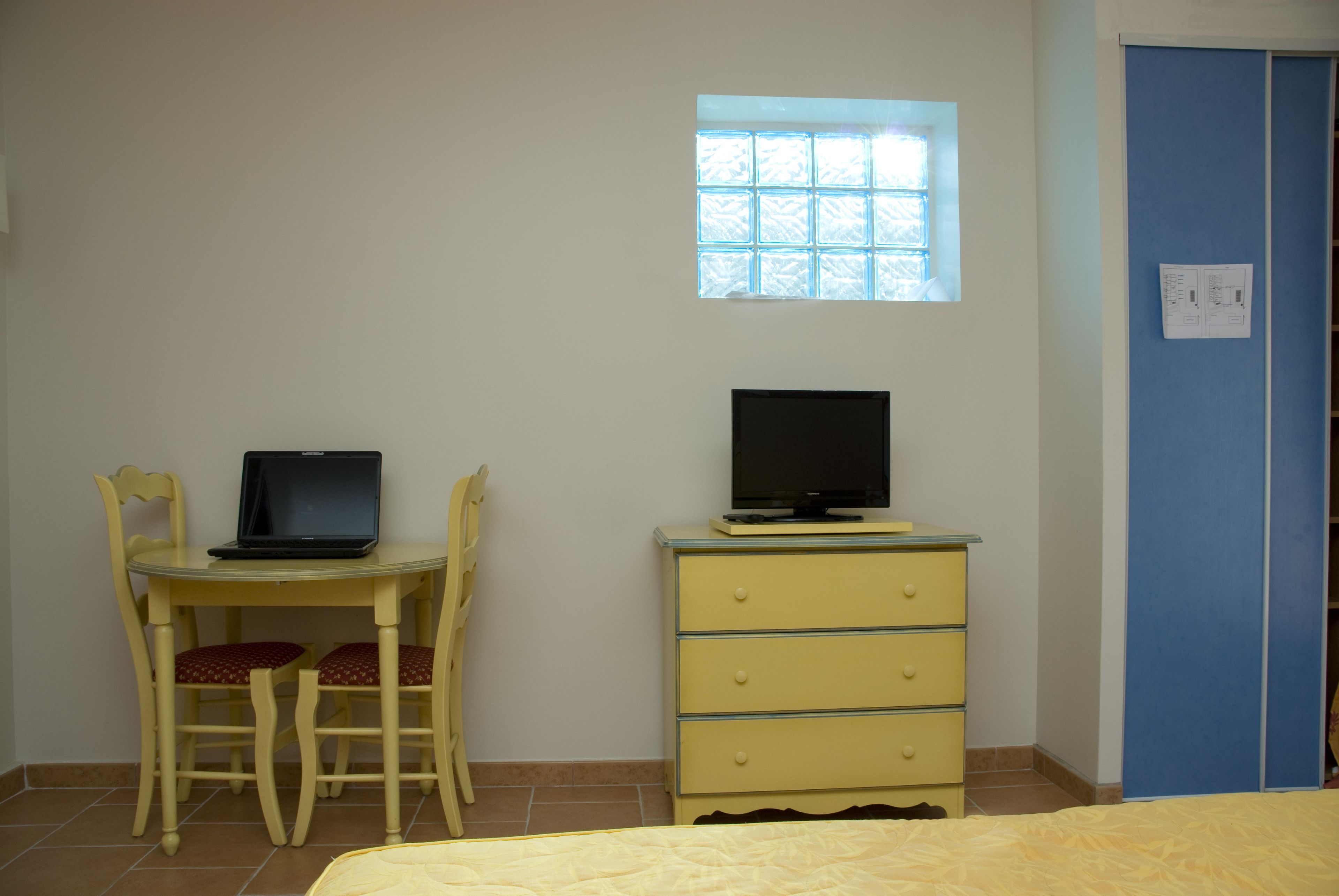 Una habitación con cama, escritorio con computadora portátil, cómoda, ventana y puerta azul.