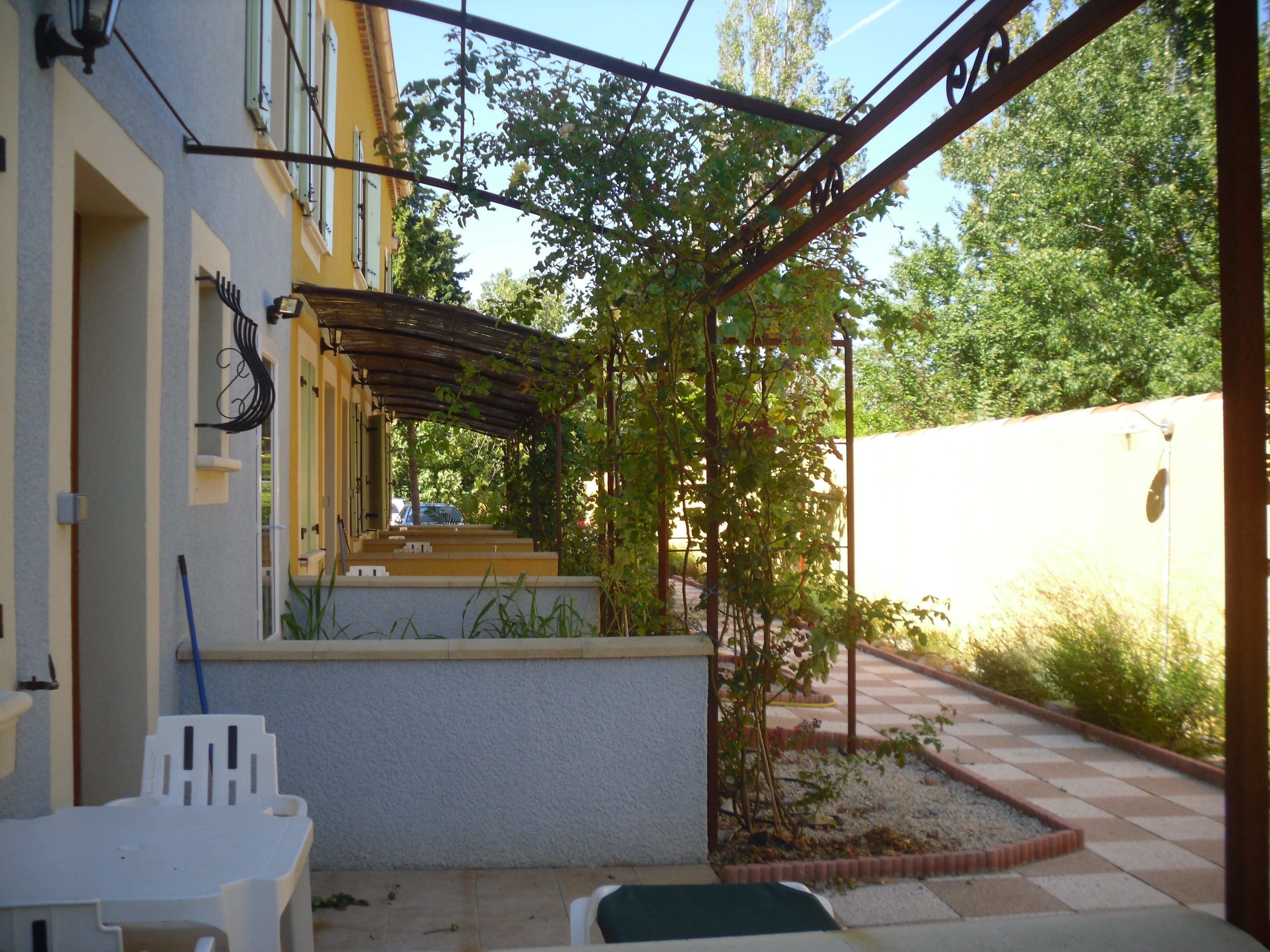 Un área de patio techada con una pérgola, una silla de plástico blanco y una mesita.