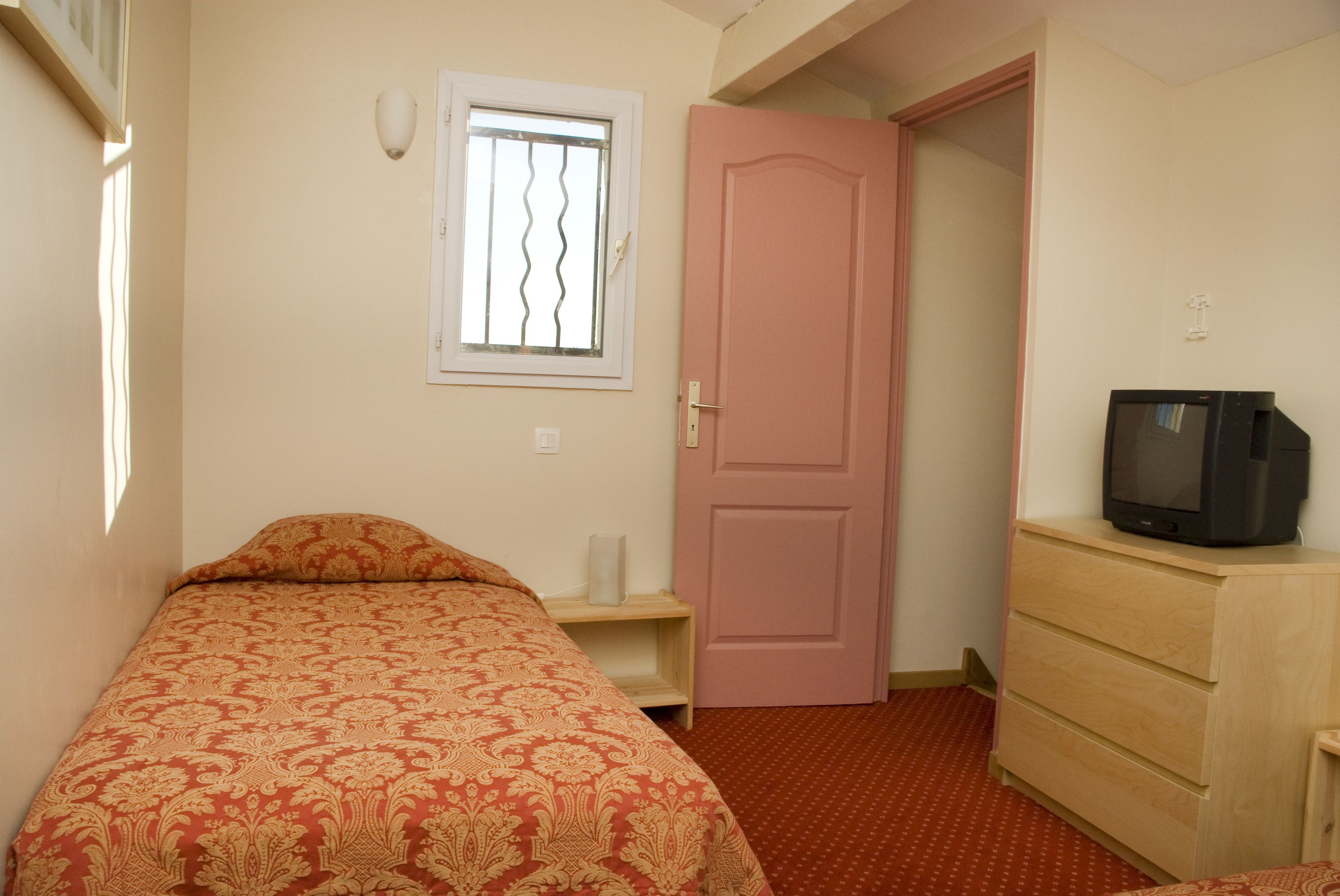 Un dormitorio con cama, cómoda, televisor, ventana con rejas y puerta.