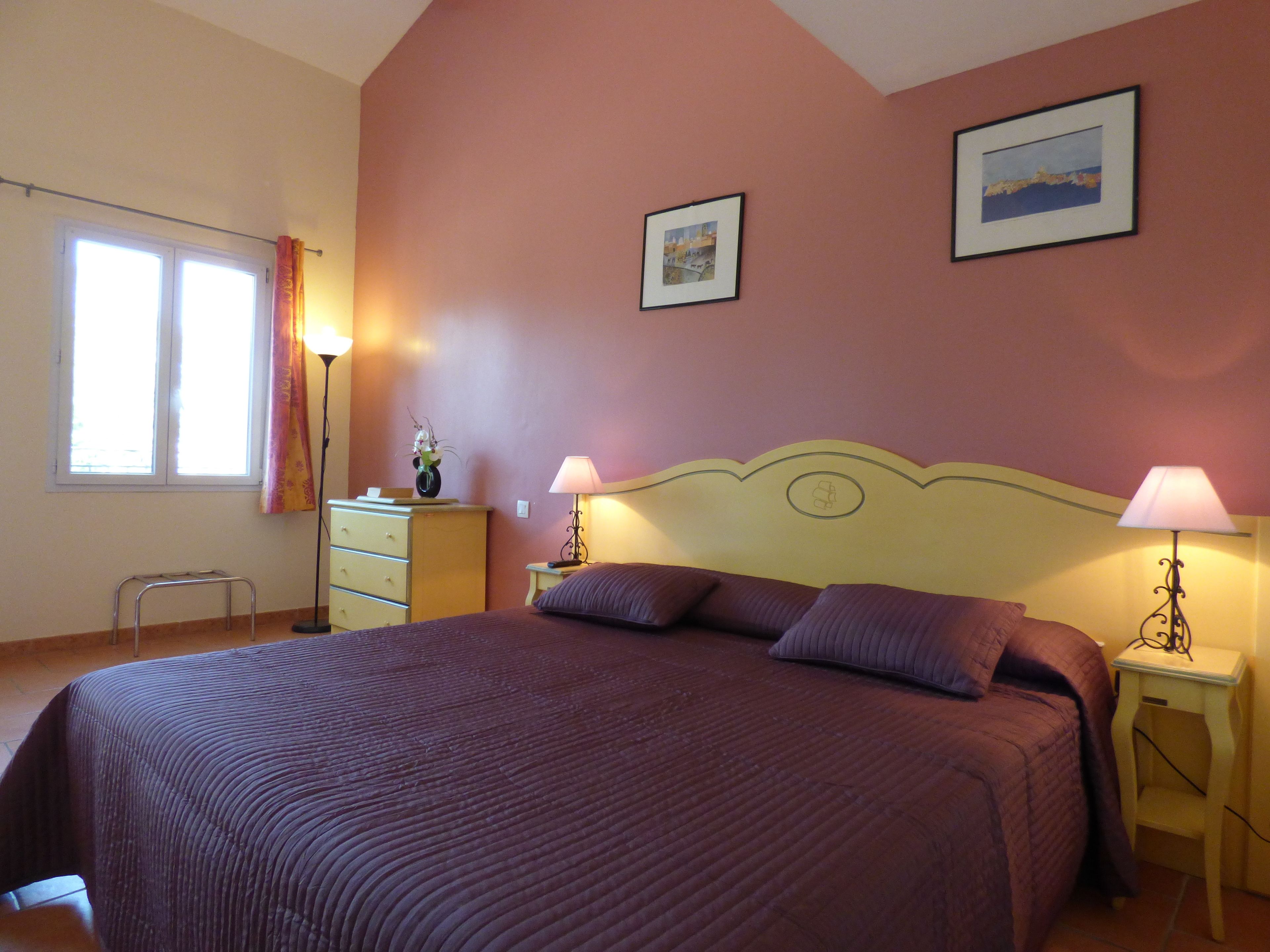 A bedroom with a large bed, two bedside lamps, a dresser, a window with curtains, and two framed pictures on the wall.