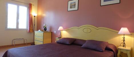 A bedroom with a large bed, two bedside lamps, a dresser, a window with curtains, and two framed pictures on the wall.