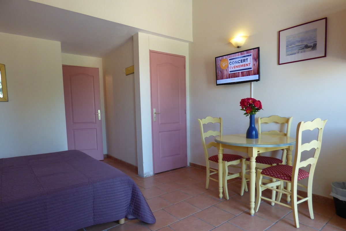 A room with a bed, a dining table with chairs, a TV on the wall, and a door leading to another room.