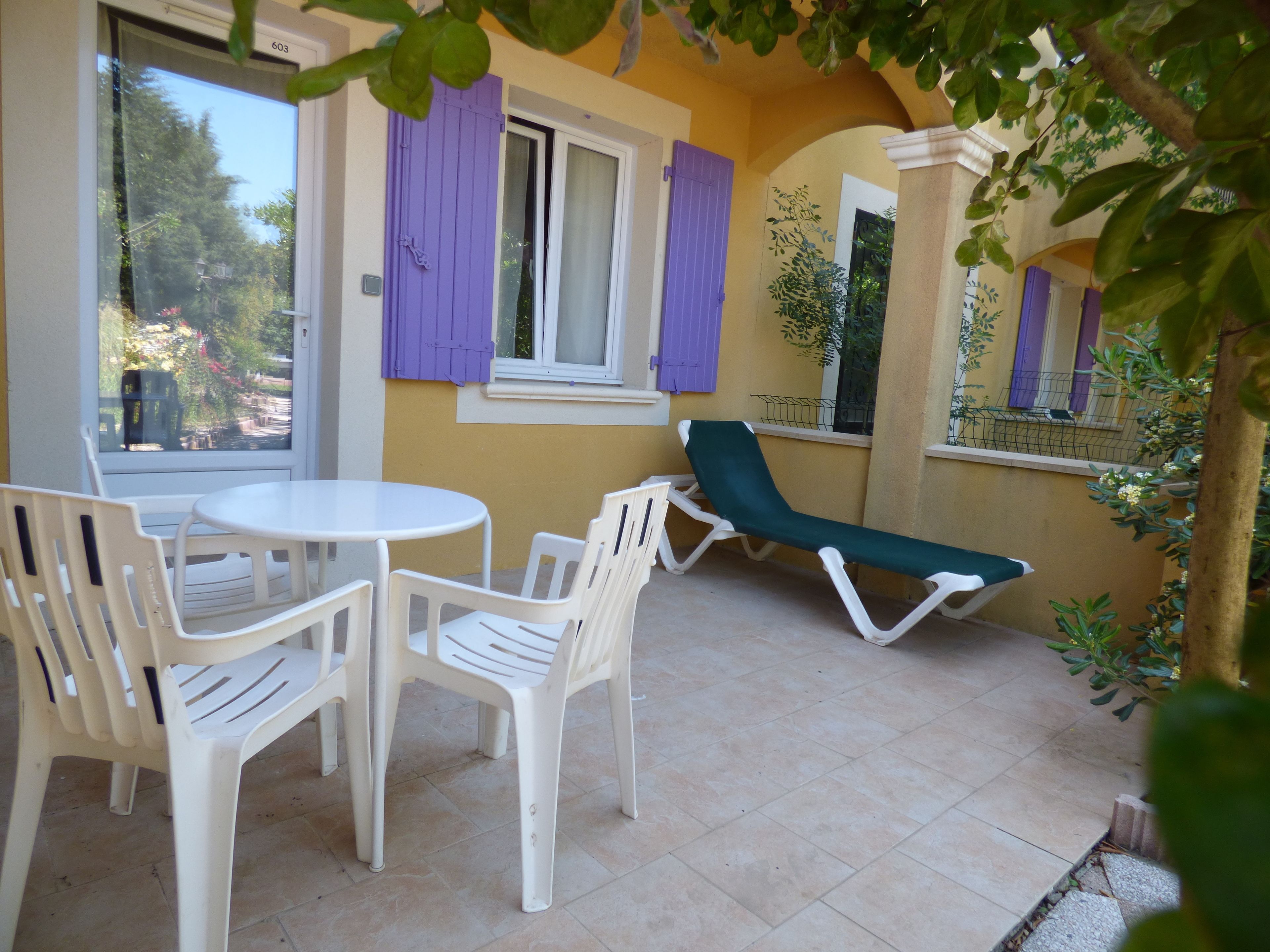 Un patio con una mesa y sillas blancas, una silla reclinable verde y una ventana con contraventanas moradas.