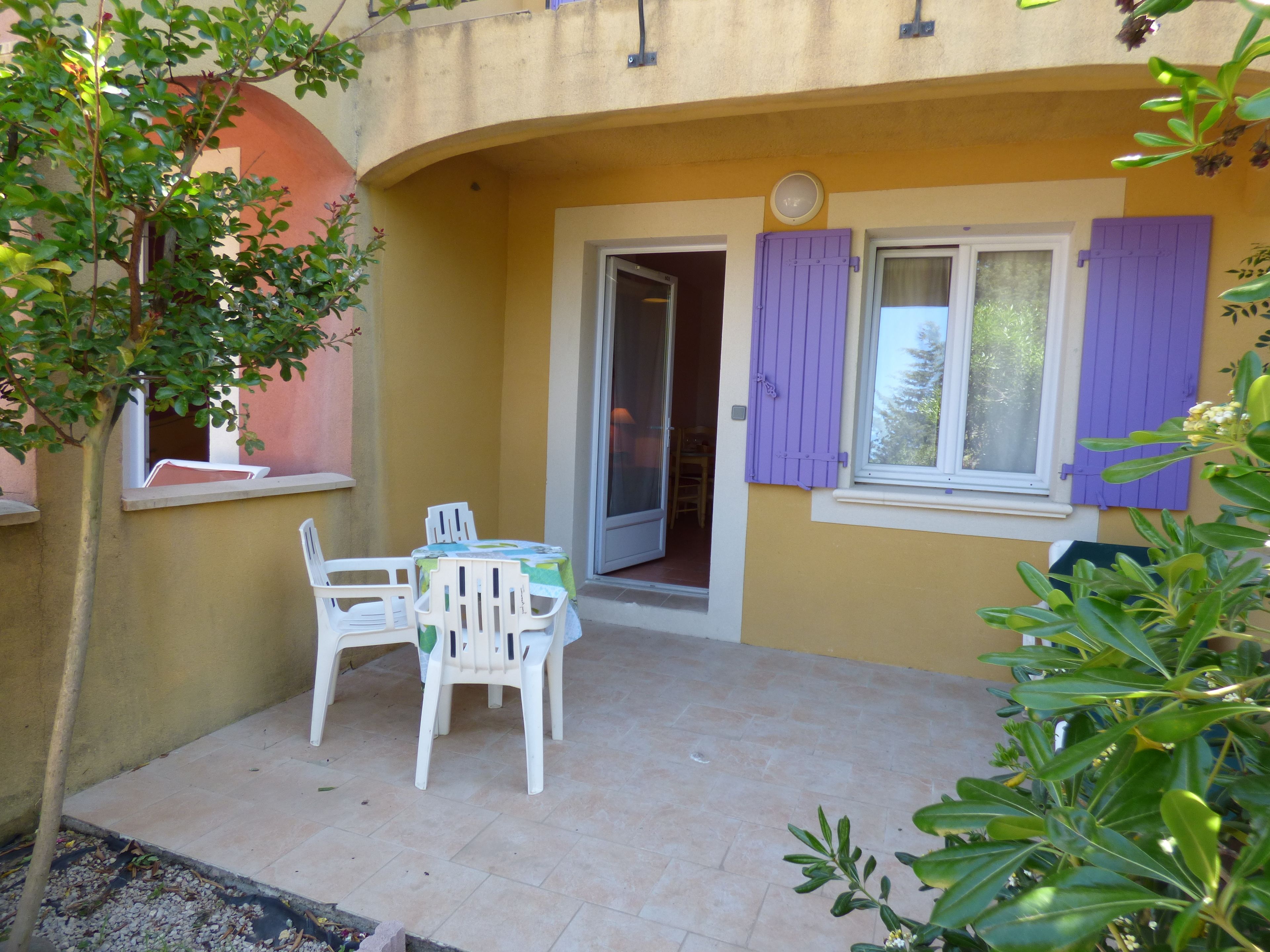 Une petite terrasse extérieure avec une table et des chaises, une porte donnant sur l’intérieur et une fenêtre avec des volets violets.