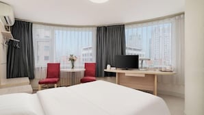 A hotel room with a large bed, a desk with a TV, two red chairs, and a view of tall buildings through large windows.