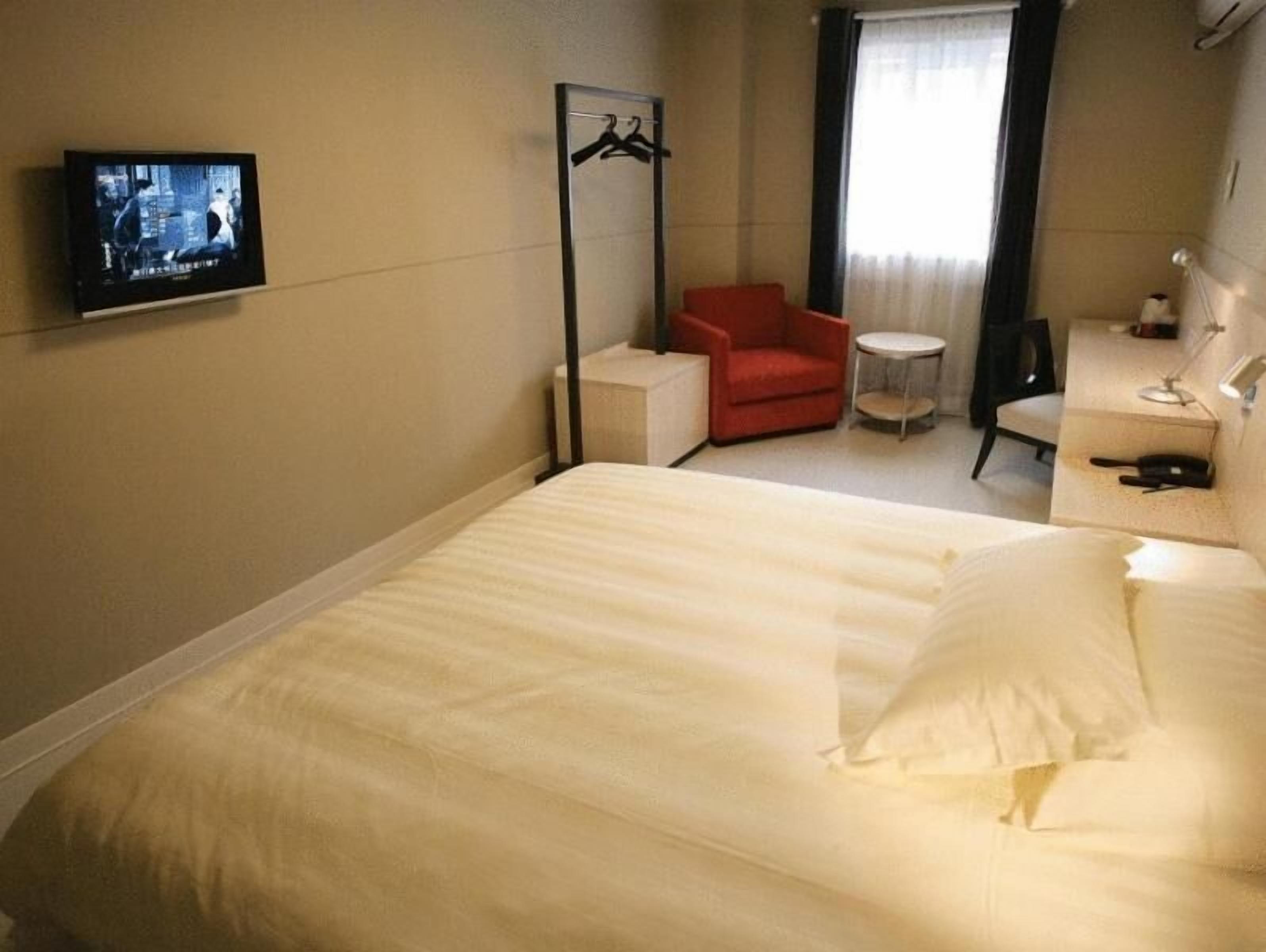 Ett hotellrum med en säng, en röd stol, ett litet bord, en tv och en klädhängare.