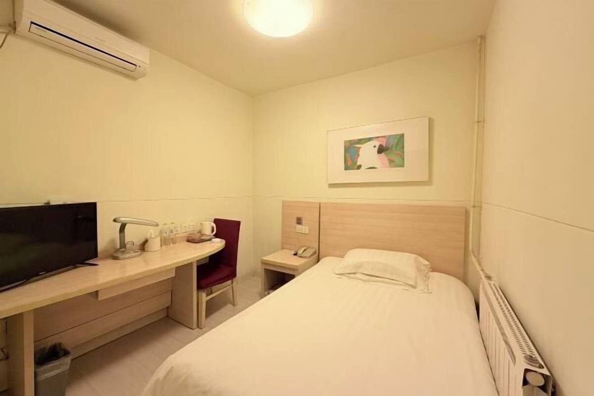 A hotel room with a bed, a desk with a TV, a chair, a bedside table, a phone, and a painting on the wall.