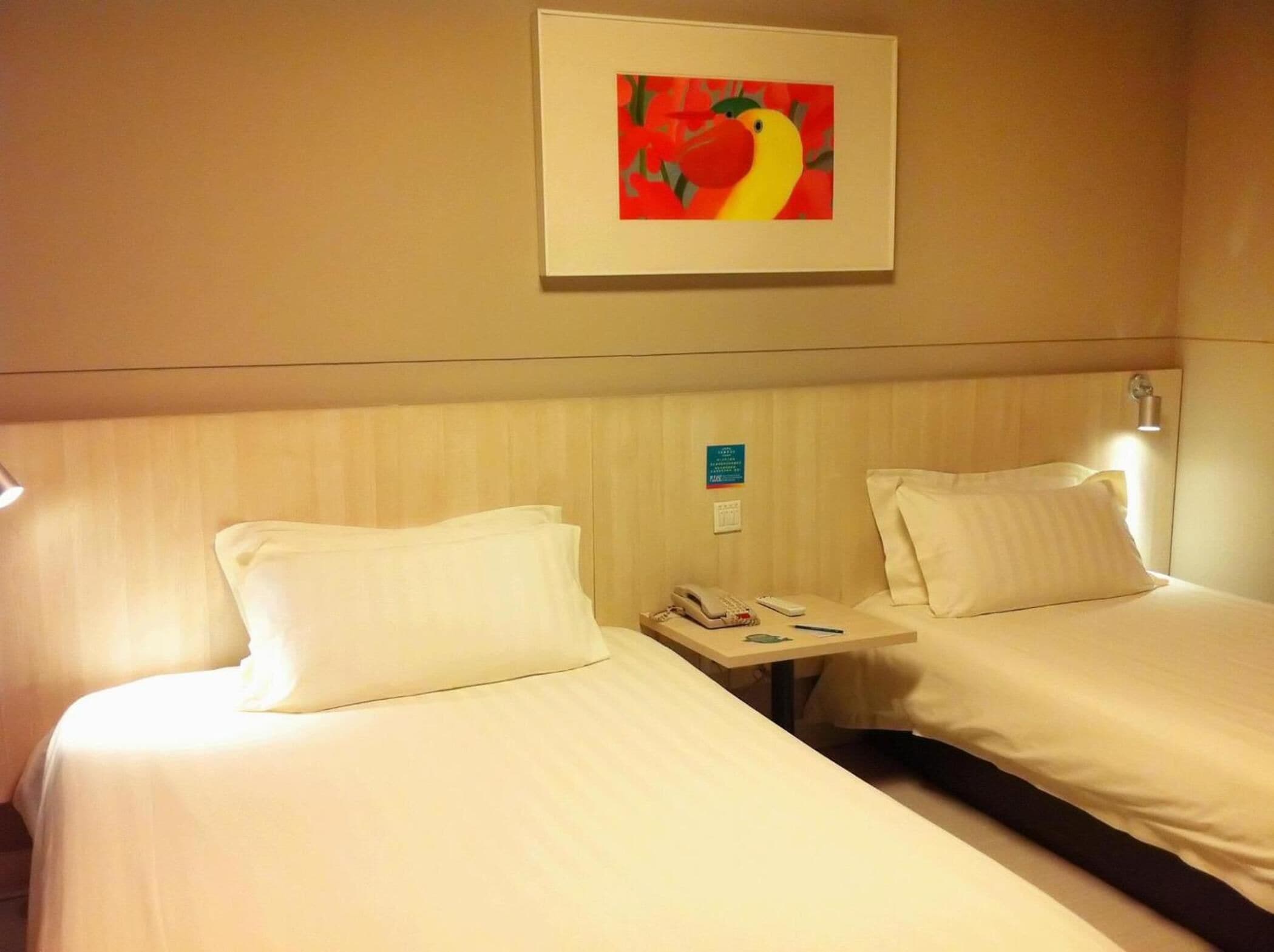 A hotel room with two beds, a bedside table with a phone and a remote control, and a framed picture on the wall.