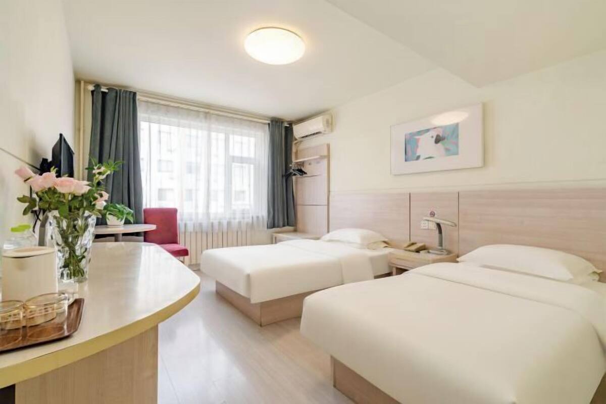A hotel room with two beds, a desk, a chair, a window with curtains, a painting on the wall, and a vase with flowers on the desk.
