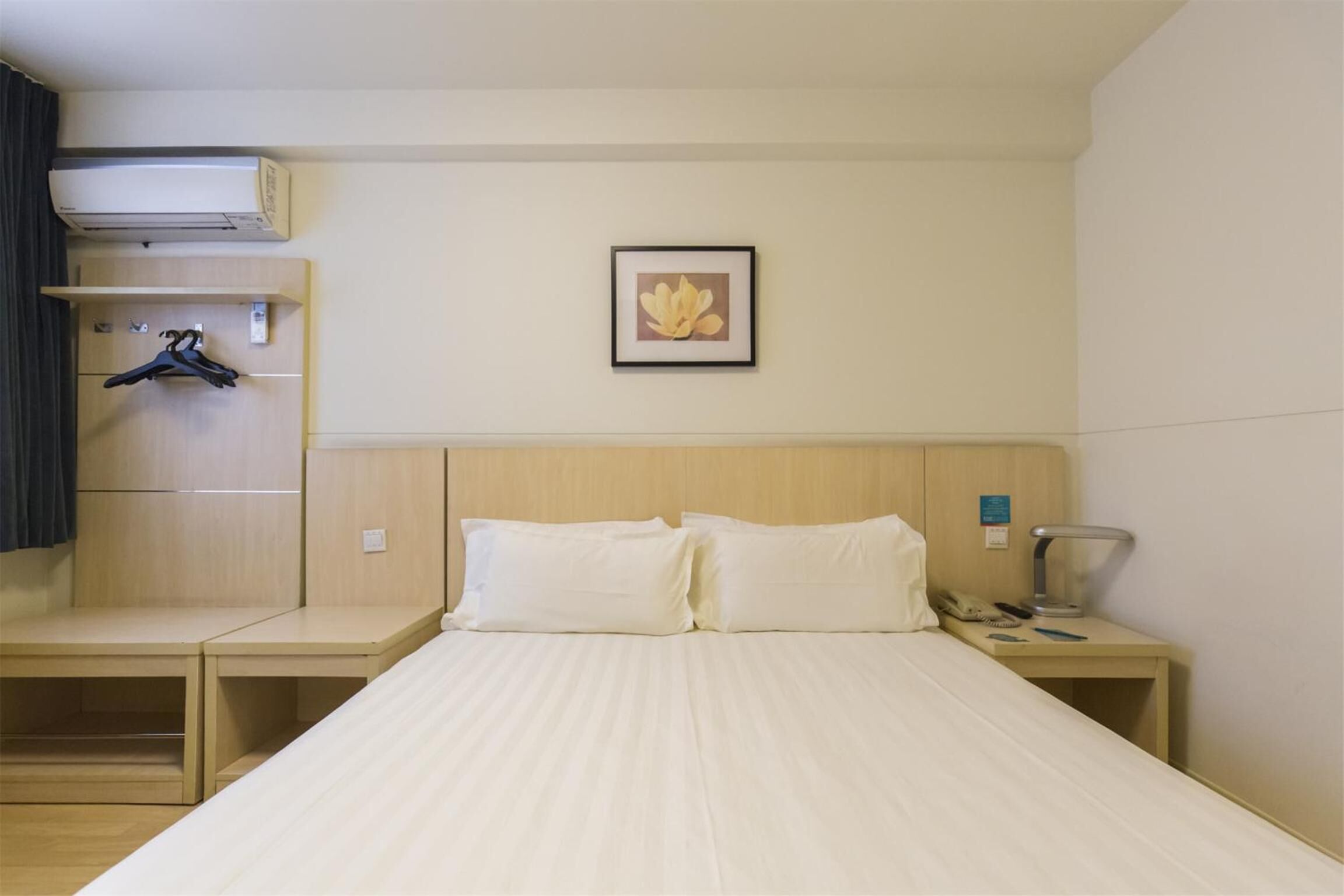 Una habitación de hotel con una cama, dos mesitas de noche, una lámpara, un cuadro enmarcado en la pared y un perchero con perchas.