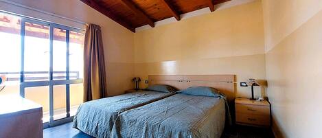 A bedroom with a wooden ceiling, a large window with curtains, two beds with matching bedding, a bedside table, and a nightstand.