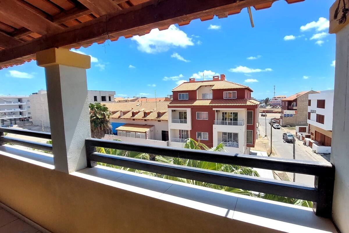 A balcony with a view of a residential area, featuring multi-story buildings with red and white facades, a clear blue sky, and a wooden pergola overhead.
