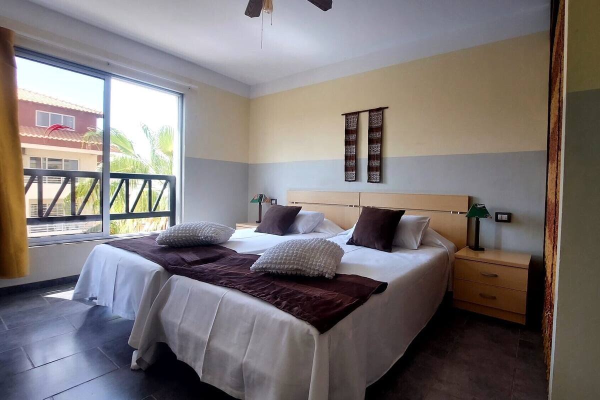 A double bed with two pillows, a nightstand, a lamp, and a window with a view of palm trees.