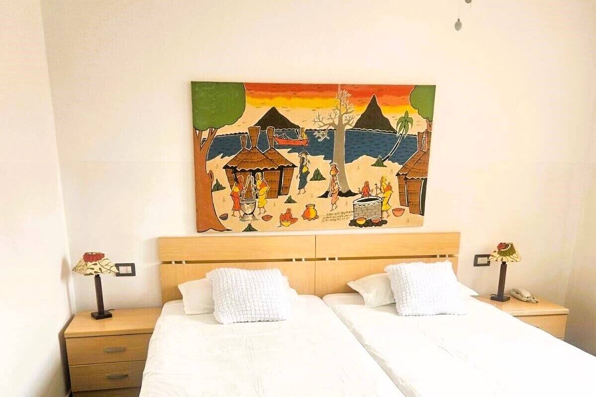 A hotel room with two single beds, a nightstand with a lamp, a painting on the wall, and a ceiling fan.