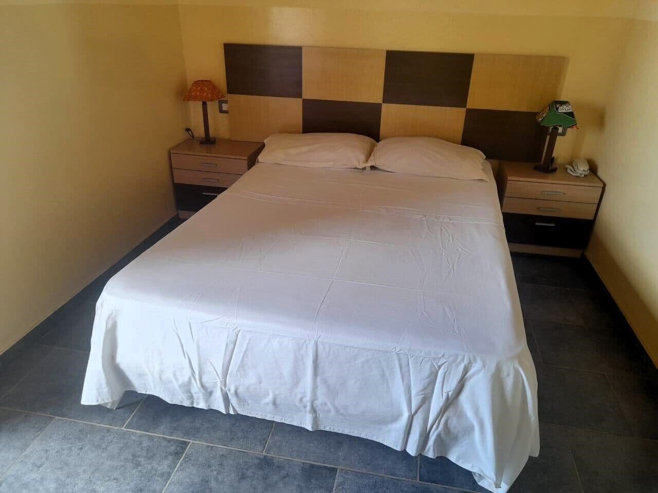 Una cama bien hecha con una colcha blanca, un cabecero a cuadros y una mesita de noche con una lámpara y un teléfono.