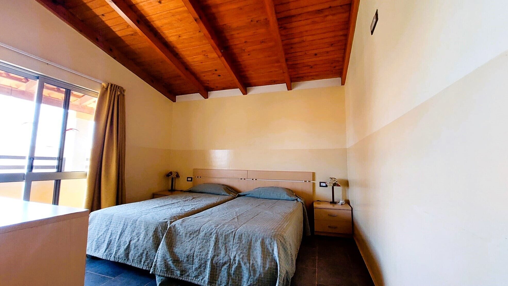 A small, clean room with two single beds, wooden ceiling beams, a window with curtains, and a bedside table with a lamp.