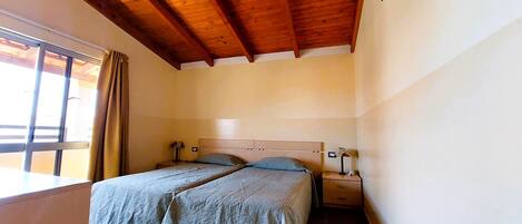 A small, clean room with two single beds, wooden ceiling beams, a window with curtains, and a bedside table with a lamp.