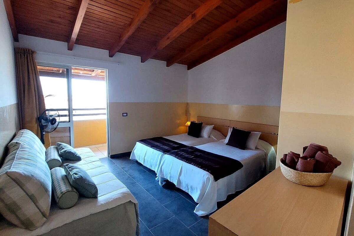 A hotel room with a bed, a sofa, a window with curtains, a ceiling fan, and a nightstand with a lamp.
