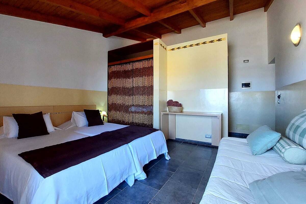 A hotel room with a large bed, a bedside table with a lamp, a small bedside table, a nightstand, a desk with a lamp, a chair, and a wall-mounted shelf.