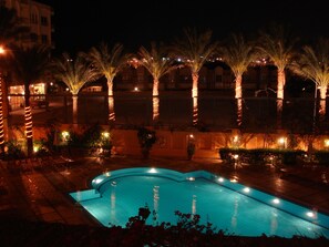 A nighttime swimming pool area with palm trees and a building in the background. - Sea Garden Hotel (Hurghada)