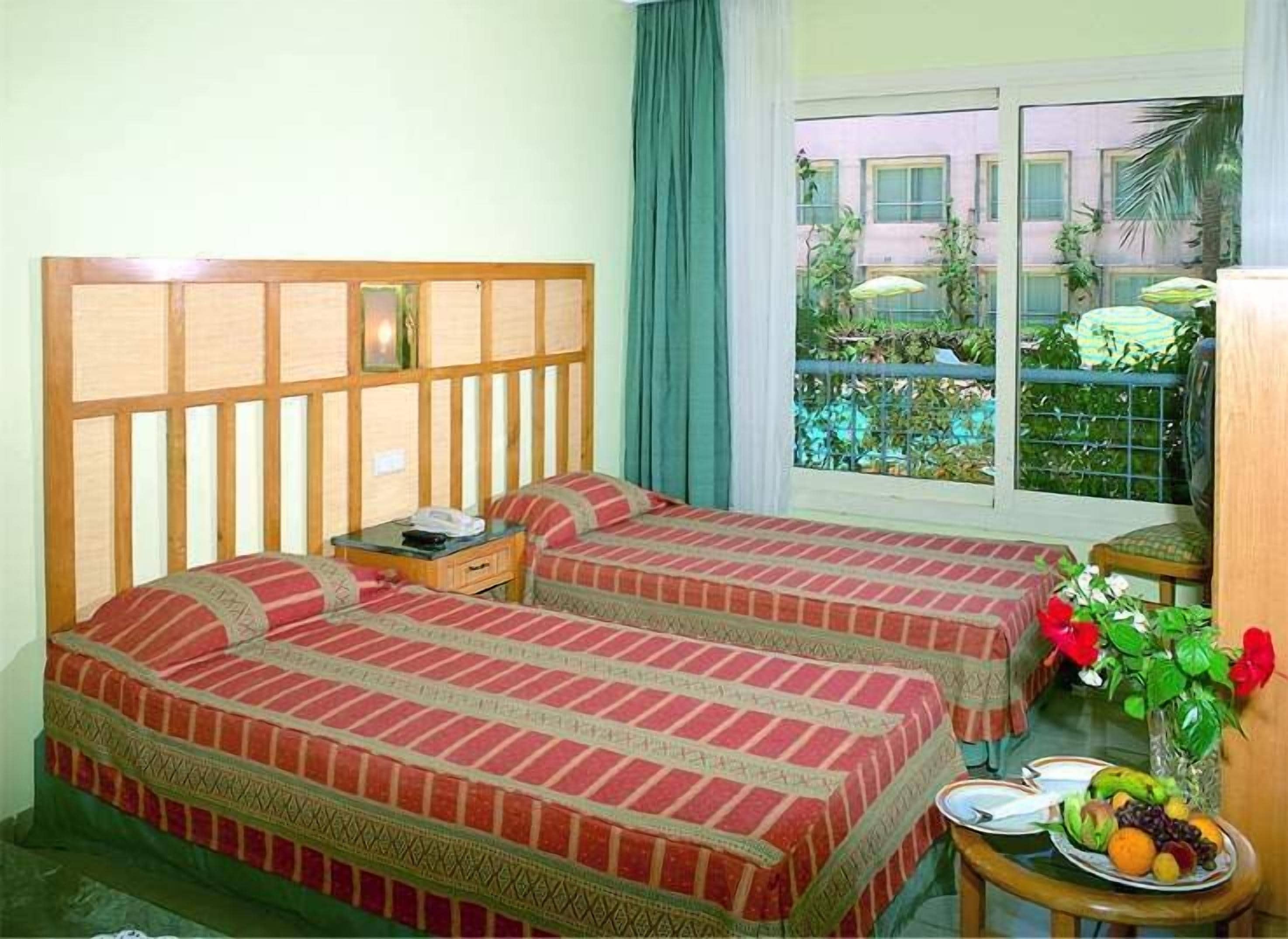 Une chambre d’hôtel avec deux lits, une petite table sur laquelle est disposée une assiette de fruits, et une vue sur un balcon agrémenté de plantes et sur un immeuble.
