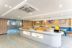 A modern hotel lobby with a central breakfast bar, seating area, and a glass partition.
