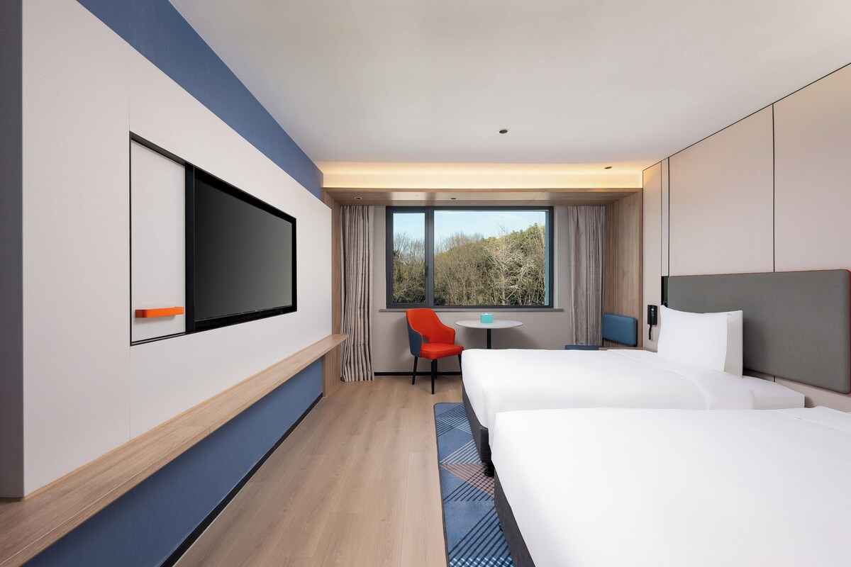 Een hotelkamer met twee bedden, een televisie, een klein tafeltje, een stoel en een raam met uitzicht op bomen.
