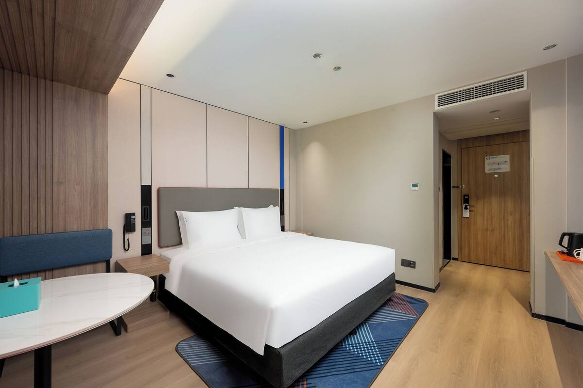 A modern hotel room with a large bed, a bedside table, a desk, and a chair.