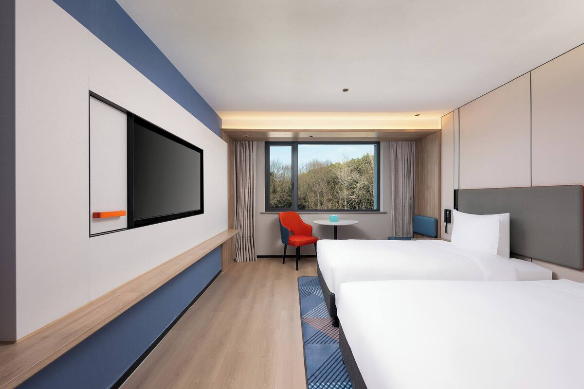 A hotel room with two beds, a TV, a small table, a chair, and a window with a view of trees.
